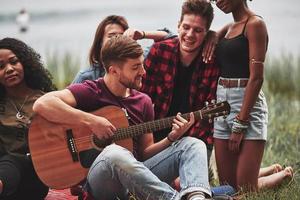 Guitarist in center of everyone's attention. Group of people have picnic on the beach. Friends have fun at weekend time photo