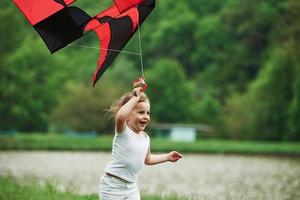 Fresh air. Positive female child running with red and black colored kite in hands outdoors photo