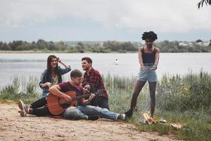 Fire is burning, music is playing. Group of people have picnic on the beach. Friends have fun at weekend time photo