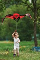 Vertical photo. Positive female child running with red and black colored kite in hands outdoors photo