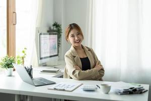 retrato de una joven y hermosa mujer de negocios asiática mirando la cámara y sonriendo, con los brazos cruzados en el lugar de trabajo de la oficina moderna foto