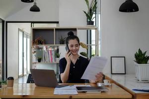 Young asian businesswoman beautiful charming smiling and talking on the mobile phone working with paperwork financial graph chart in the office photo