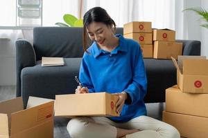 Shipping shopping online, startup small business owner writing address on cardboard box at workplace. Freelance Asian woman small business entrepreneur SME working with box at home photo