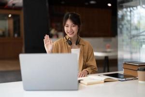 Joyful young asian female in wireless headphones waving wand greeting to screen while sitting at table and having video chat with business partners using laptop against of comfortable office photo