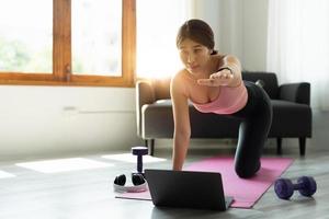 mujer asiática deportiva saludable en ropa deportiva en casa usando una computadora portátil para compartir resultados de entrenamiento en las redes sociales y haciendo ejercicios abdominales en la alfombra de fitness foto