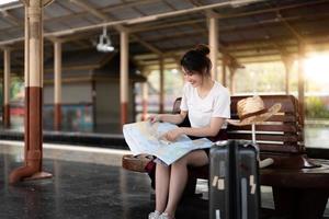 Happy young asian woman traveler or backpacker using map choose where to travel with luggage at train station, summer vacation travel concept photo