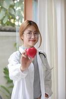 retrato joven médico asiático hermoso con estetoscopio sosteniendo el corazón mientras está de pie en su oficina.