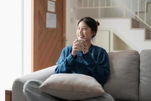 feliz joven asiática sentada en un sofá con una taza de café en casa foto