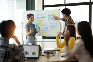 group of young creative asian people doing brainstorming meeting colleagues in board room discussing project. woman standing at white board give presentation photo