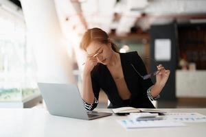 Very stressed business asian woman sitting in front of her computer looking at a large pile of paperwork, while holding a hand at her forehead photo
