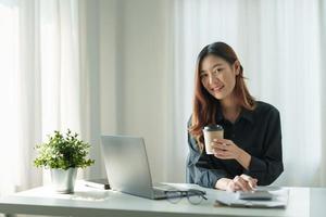 Charming asian female office worker working on a laptop computer and enjoying drinking coffee in a modern office. photo