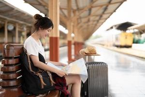 young asian woman traveler sitting with map choose where to travel and bag waiting for train at train station, summer vacation travel concept photo