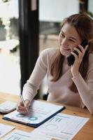 Portrait of young asian woman sitting at her desk working with calculator and paperwork while calling mobile phone in modern office. photo