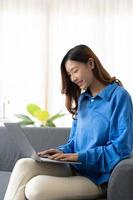 Young asian business woman smile and work from home with laptop computer online to internet on sofa in living room, freelance girl using notebook on couch with comfort, new normal, lifestyle concept photo