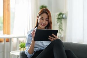 Charming asian woman using digital tablet for online shopping chatting on sofa at living room. photo