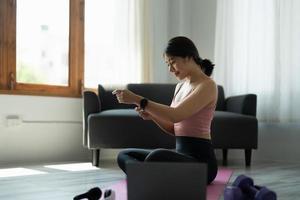 Attractive young asian fitness woman  in sportswear resting after workout using smartwatch  for online training at home photo