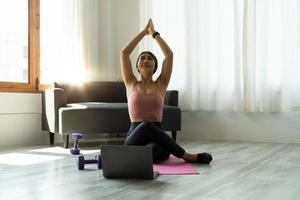 tranquila y relajada mujer asiática joven sentada en posición de loto con las manos en alto gesto de namaste, ojos cerrados, respirando, descansando, meditando profundamente, practicando yoga en la alfombra de yoga en casa foto