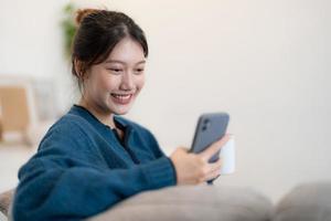 feliz joven asiática usando teléfono móvil mientras se sienta en un sofá en casa foto
