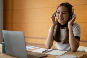 una estudiante asiática sonriente usa audífonos inalámbricos para estudiar en línea, una joven feliz aprende un idioma escucha una conferencia mira un seminario web escribe notas mira una laptop en casa, educación distante foto