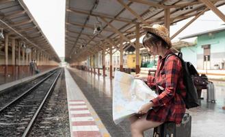Young woman traveler with backpack looking to map while waiting for train, Asian backpacker on railway platform at train station. Holiday, journey, trip and summer summer travel concept photo