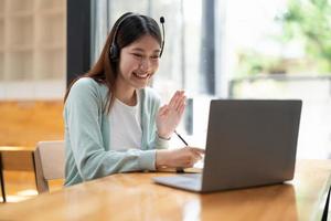 Smiling girl student wear wireless headphone study online with teacher, happy young asian woman learn language listen lecture watch webinar write notes look at laptop sit in cafe, distant education photo