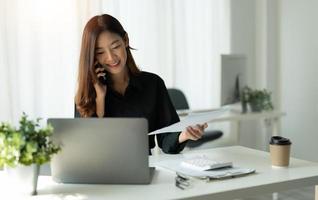 Young Asian Business Woman Using Computer in office with notebook and calling on mobile phone, financial accounting concept. photo
