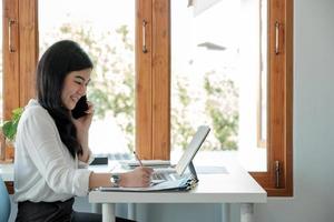 vista lateral feliz mujer de negocios asiática sentada en su lugar de trabajo en la oficina. mujer joven que trabaja para gráficos de datos de documentos financieros con una computadora portátil durante una llamada telefónica en la oficina foto
