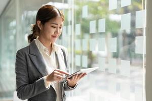 atractiva mujer asiática de negocios parada cerca de la ventana y usando una tableta digital inteligente. foto