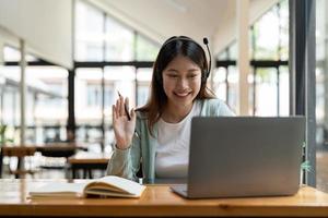 Attractive happy young student studying online at home, using laptop computer, headphones, having video chat, waving. Remote work, distance education. Video conference or virtual event on quarantine photo