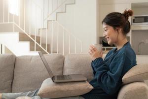una joven asiática tranquila que trabaja en una laptop sentada en un sofá acogedor, una chica feliz navegando por Internet o comprando en línea durante el fin de semana soleado en casa, relajándose en el sofá con la computadora foto