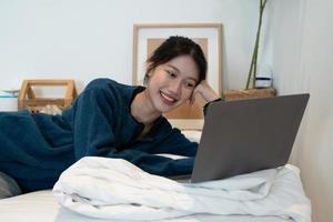 encantadora chica asiática disfrutando de una película en su computadora portátil. linda adolescente viendo videos en línea en la cama foto