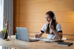 una estudiante sonriente usa notas para estudiar en línea con el maestro, una joven feliz aprende el idioma escucha una conferencia mira un seminario web escribe notas mira una laptop en casa, educación distante foto