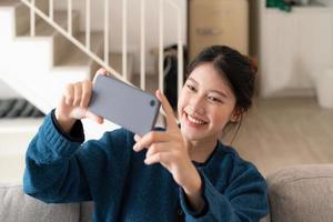 Photo of young asian woman taking selfie on cell phone while sitting on couch in bright apartment