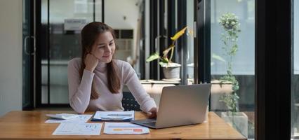 joven empresaria asiática sonriente que usa una computadora en el lugar de trabajo de la oficina en casa, una feliz empleada coreana que trabaja en una laptop, una estudiante atractiva que estudia comunicarse en línea con una computadora portátil. foto