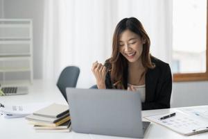 Happy young asian business woman in suit looking at laptop excited by good news online, lucky successful winner man sitting at office desk raising hand in yes gesture celebrating photo