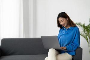 joven mujer de negocios asiática sonríe y trabaja desde casa con una computadora portátil en línea a Internet en un sofá en la sala de estar, una chica independiente que usa un cuaderno en el sofá con comodidad, nuevo concepto de estilo de vida normal