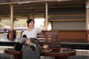 Happy young asian woman traveler or backpacker using map choose where to travel with luggage at train station, summer vacation travel concept photo
