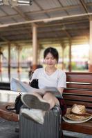 Happy young asian woman traveler or backpacker using map choose where to travel with luggage at train station, summer vacation travel concept photo