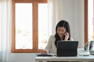 feliz gerente asiática de negocios trabajando en su escritorio en la oficina tomando una llamada en su teléfono móvil mientras escribe notas en un bloc de notas foto
