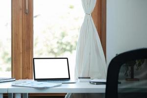 Mockup laptop with blank screen on white table with calculator and graph chart paperwork. Home interior or office background photo