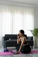 retrata a una mujer deportiva asiática con ropa deportiva haciendo ejercicio y usando una laptop y llamando por teléfono a casa en la sala de estar, sentada en el suelo con pesas en una alfombra de yoga. concepto de deporte y entrenamiento en línea foto