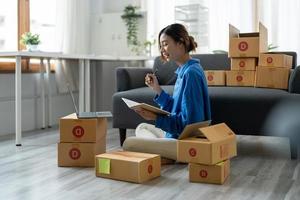 Sell online, Young asian woman checking order and customer's details and addresses on laptop or box in order to prepare for shipping according to the information, Online shopping SME entrepreneur photo