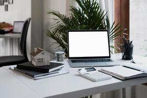 computadora portátil con pantalla blanca en blanco en un escritorio moderno, bienes raíces con modelo de casa, bloc de notas y calculadora en el lugar de trabajo foto