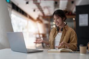 mujer asiática sonriente independiente con auriculares, comunicándose con el cliente a través de una videollamada. tutora profesional milenaria y agradable que da clases de idiomas en línea. foto