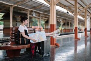 Happy young asian woman traveler or backpacker using map choose where to travel with luggage at train station, summer vacation travel concept photo