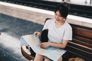 feliz joven mujer asiática viajera o mochilera que usa el mapa elige dónde viajar con equipaje en la estación de tren, concepto de viaje de vacaciones de verano foto