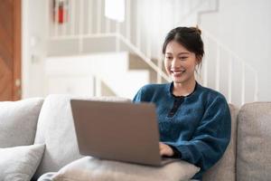 feliz casual hermosa mujer asiática joven que trabaja en una computadora portátil sentada en el sofá de la casa. foto