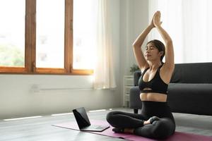 tranquila y relajada mujer asiática joven sentada en posición de loto con las manos en alto gesto de namaste, ojos cerrados, respirando, descansando, meditando profundamente, practicando yoga en la alfombra de yoga en casa. foto