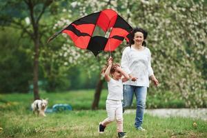 Weekend activities. Positive female child and grandmother running with red and black colored kite in hands outdoors photo