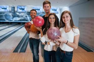 Standing against playing area. Young cheerful friends have fun in bowling club at their weekends photo